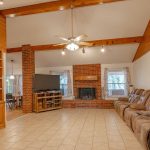 A spacious living room with a tiled floor, brown recliner sofas along one wall, a brick fireplace, a TV on a stand, built-in wooden shelves, and a ceiling fan with lights hanging from a wood-beamed ceiling.