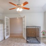 A bright room with a ceiling fan, French doors, a large dog crate, a white dresser, houseplants on a red cooler, and a window with curtains letting in natural light.