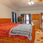 A cozy bedroom with wooden furniture, including a bed with storage drawers, a dresser, a nightstand, and an armoire. The bed has a red bedspread and a blue-gray blanket. A ceiling fan and natural light brighten the room.