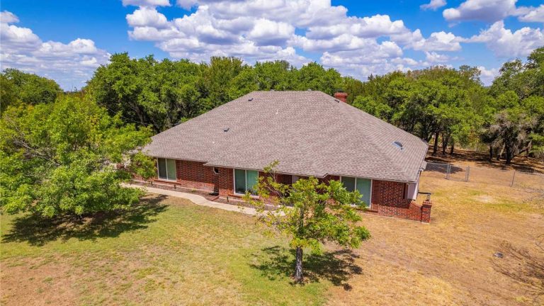 A single-story brick house with a large gray shingle roof sits in a spacious yard with patches of grass and a few trees, surrounded by a wooded area under a blue sky with scattered clouds.