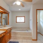 A spacious bathroom with double sinks, wooden cabinets, a large mirror, a built-in bathtub with marble surround, a ceiling fan, and a window with decorative glass. A separate toilet area is visible on the right.