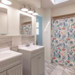 Bright bathroom with two white sinks and mirrors, light gray tiles, and a shower with a colorful floral curtain. Overhead lighting and a skylight brighten the space. Marble-pattern floor tiles complete the room.