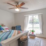 A cozy bedroom with a ceiling fan, bed with a colorful striped blanket and stuffed animals, a desk with a blue chair, a window with curtains, and various furniture and decor items. Sunlight shines through the window.