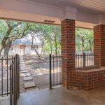 A covered patio with brick columns and iron gates overlooks a yard with stepping stones, trees, and a building in the background. A black grill sits on the right side of the patio.
