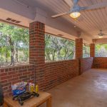 A covered patio with red brick walls, a ceiling with two ceiling fans, and a concrete floor. There is a small grill and condiments on a wooden table, with trees and a yard visible through open windows.