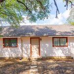 A small, single-story house with white walls, a brown roof, and red trim sits among trees. There are two windows, a central door, a path of stepping stones, and a white chair on the right side near the entrance.