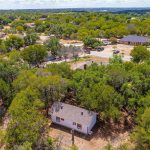 A small white house with a brown roof sits surrounded by trees in a semi-rural neighborhood. Other homes, roads, and green spaces are visible in the background under a partly cloudy sky.