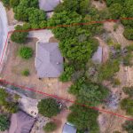 Aerial view of a house surrounded by trees, with the property boundary outlined in red. The lot is triangular and bordered by a road on one side, with sparse grass and neighboring homes nearby.