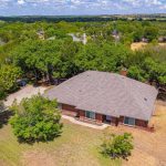 Aerial view of a single-story brick house with a gray roof, surrounded by trees and grassy yard, set in a spacious rural neighborhood under a partly cloudy sky.