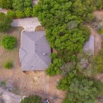 Aerial view of a house with a gray roof surrounded by trees and dry grass. A driveway leads to the road on the left, and another structure is partially visible through the trees.