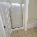 A bathroom with beige tile flooring, a glass-enclosed shower with a chrome frame, a white panel door, and a partial view of a white bathtub. The walls are painted off-white.