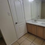 A bathroom with beige tile floor, a white door, a large mirror above a countertop with a sink, wooden cabinets below, and bright overhead lights.