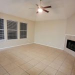 A spacious, empty living room with beige tiled floors, light-colored walls, three large windows with blinds, a ceiling fan with lights, and a corner fireplace with a white tile surround.