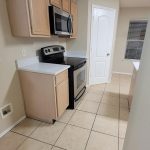 A kitchen with light wood cabinets, a stainless steel oven and microwave, beige tiled floor, white countertops, and a white closed door at the end of the room. A window with blinds is on the right wall.