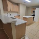 Modern kitchen with light wood cabinets, white countertops, a center island, stainless steel sink, electric stove, and beige tiled floor under bright overhead lighting.