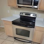A kitchen with tan cabinets, a stainless steel electric stove with a glass cooktop, and a matching microwave above it. The countertops are white, and the floor is covered in beige tiles.