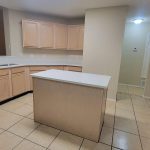A modern kitchen with light wood cabinets, white countertops, a stainless steel dishwasher and oven, a central island, tile flooring, and neutral-colored walls. The space is well-lit with overhead lighting.