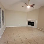 A spacious, empty living room with beige tiled flooring, light-colored walls, a ceiling fan with light, a corner fireplace with white tile, and three large windows with blinds.