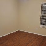 An empty room with beige walls, wood flooring, a ceiling fan with lights, and a window with blinds partially open.