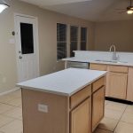 A modern kitchen with light wood cabinets, white countertops, a center island, stainless steel sink, white appliances, large windows with blinds, tile flooring, and a back door. Ceiling lights are on.