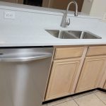A clean kitchen countertop with a stainless steel sink, a silver dishwasher, light wood cabinets, and beige tiled floor. Multiple electrical outlets are visible on the wall above the counter.