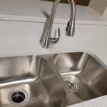 A modern double-basin stainless steel kitchen sink with a curved faucet, set in a speckled white countertop. An electrical outlet is visible on the right wall above the counter.