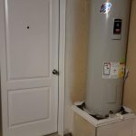 A white residential door is next to a tall, cylindrical water heater mounted on a platform in a garage or utility room with unfinished walls and a bare concrete floor.