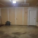 A mostly empty garage with unfinished walls, a concrete floor, a white door at the back, a water heater on the right, shelves with items, and a gray storage container and broom on the left.