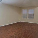 Empty room with wood flooring, beige walls, white trim, and two windows. A ceiling light hangs on the left, and a white entry door with tile flooring is on the right.