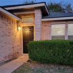 Single-story brick house at dusk with a black front door and lit porch light, two windows with blinds, and a neatly trimmed hedge in front. Trees and a blue evening sky are visible in the background.
