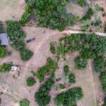 Aerial view of a rural landscape near De Leon TX, with scattered trees, a house, vehicles, open grassy areas, a dirt road along County Road 419, and a small pond bordered by dense greenery.