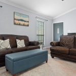 A living room with two brown leather sofas, a blue upholstered ottoman, light walls, large windows with blinds, and a landscape painting above the sofa. A door and a rug are also visible.
