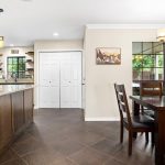 A modern kitchen and dining area with dark wood cabinets, granite countertops, stainless steel appliances, and a wooden dining table with six chairs under a contemporary chandelier. A window and artwork are on the cream-colored wall.
