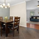 Dining area with a dark wooden table and six chairs, a flower vase centerpiece, tile flooring, and a modern chandelier. In the background, a living room with sofas, a ceiling fan, and large windows is visible.