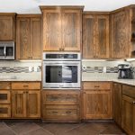 A kitchen with wooden cabinets, a built-in microwave and oven, black and white checkered backsplash, granite countertops, a coffee maker, a knife block, and a window above the sink.