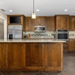 Modern kitchen with wood cabinets, stainless steel appliances, granite countertops, tile backsplash, and pendant lights over an island. A window and sink are on the right, and the floor is dark tile.