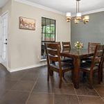 A modern dining room with dark wood table and six chairs, a small flower arrangement, a gray accent wall, tile flooring, a ceiling light fixture, a window, white closet doors, and mountain artwork on the wall.
