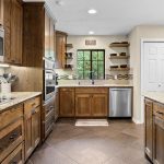 Modern kitchen with wooden cabinets, granite countertops, stainless steel appliances, a window above the sink, open shelves, and a large island. Neutral tile flooring and double doors are visible in the background.