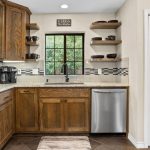 A kitchen with wooden cabinets, stainless steel oven and dishwasher, floating shelves with black dishes, a coffee maker, a window above the sink, and a rug on the tiled floor. A sign above the window reads, "Alexa, do the dishes.