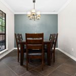 A modern dining room with a dark wooden table and six matching chairs, a light fixture above, tiled floor, a blue accent wall, and a mountain landscape painting on a white wall. A window shows greenery outside.