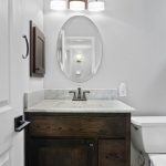 A modern bathroom with a marble-top vanity, dark wood cabinet, oval mirror, wall-mounted light, white toilet, and a bathtub with a glass shower door and gray tile.