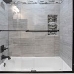 Modern bathtub and shower with glass sliding doors, gray tiled walls, black fixtures, and a recessed shelf with mosaic tile accent. Part of a white toilet is visible on the left side.