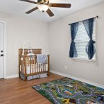 A simple nursery with a crib, a ceiling fan, blue curtains on a window, hardwood floor, and a colorful play mat with roads and buildings. The walls are light beige, and a white door is closed.