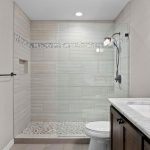 Modern bathroom with a large walk-in shower featuring a glass wall, pebble stone shower floor, tiled walls with decorative strip, a toilet, and a vanity with a marble countertop and mirror.