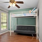 A bedroom with wood flooring features a white metal loft bed above a dark gray futon, green accent wall, window with blinds, and ceiling fan with lights.