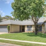 A single-story brick house with a gray roof and attached two-car garage, surrounded by a well-kept lawn and trees, sits on a quiet residential street under a partly cloudy sky.