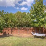 A backyard with green grass, a wooden fence, leafy trees, and a gray hammock on a metal stand under a partly cloudy sky.
