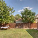 A backyard with green grass, a wooden fence, trees, a hammock on a metal stand, and part of a brick house with a covered patio on a sunny day.