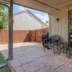 A covered patio with two wicker chairs, a small glass table, and a grill next to a wooden fence. The patio overlooks a small yard with a fire pit and some trees.