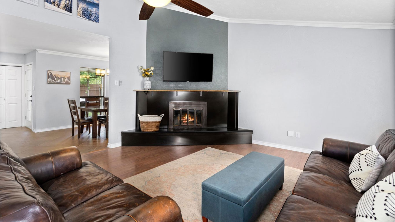 A cozy living room with brown leather sofas, a blue ottoman, a fireplace with a TV above it, hardwood floors, and a dining area visible through an open doorway.